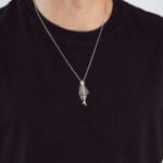 A man in a black shirt showcases a silver fish pendant with chain, embodying elegance and oceanic charm in his accessory choice.